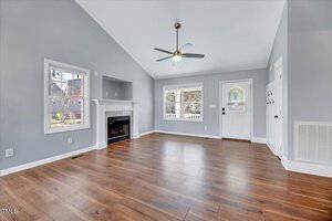 A spacious living room with polished wood flooring, gray walls, and a vaulted ceiling. It features a ceiling fan, a fireplace with a white mantel, large windows, and a white door, creating a bright and airy atmosphere.