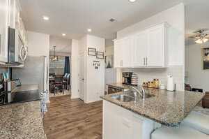 Modern kitchen with granite countertops, stainless steel appliances, and white cabinets. A breakfast bar has two chairs. The room leads to a dining area with a wooden table and large window. Warm wood floors extend throughout the open space.