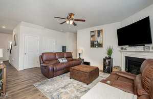 A cozy living room with brown leather sofas, a large ottoman, and a flat-screen TV mounted above a fireplace. The walls are adorned with modern artwork, and a ceiling fan hangs above. Wooden floors and natural light complete the inviting space.