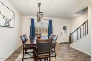 Dining room with a dark wooden table and six chairs, a large window with blue curtains, and a picture of a highland cow on the wall. The room has hardwood floors and is adjacent to an entrance door with a decorative glass pane.