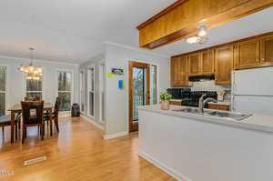 A kitchen and dining area with light wood floors. The kitchen features wooden cabinets, a white refrigerator, and a sink on an island. The dining area has a table with six chairs near large windows and a door.