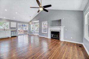 A spacious living area with light gray walls and hardwood floors. The room features a ceiling fan, a white fireplace, and large windows. An open kitchen with white cabinets and a dining space with a hanging light fixture is visible.