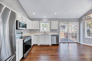 A modern kitchen with stainless steel appliances, including a refrigerator, stove, oven, and dishwasher. White cabinets and a double sink are positioned beneath large windows. The room features wooden flooring and sliding glass doors leading to a deck.