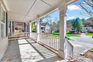 A spacious porch with white railings overlooks a suburban neighborhood. Trees, grass, and several houses are visible under a bright blue sky with scattered clouds. It's a sunny day, casting shadows on the porch floor.