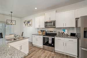 Modern kitchen with white cabinets, stainless steel appliances, and a granite countertop. A sink island is in the foreground, and a wall map decorates the background. Hardwood floors and a hanging light fixture complete the space.