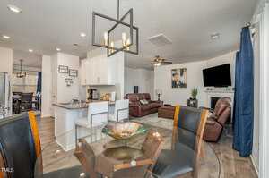 A modern open-concept living space with a dining area featuring a glass table and dark chairs, a kitchen with white cabinets and bar stools, and a living room with brown leather sofas and a wall-mounted TV above a fireplace. A stylish light fixture hangs above.
