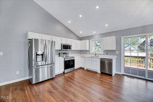 Modern kitchen with stainless steel appliances, white cabinets, wooden floors, and gray walls. Includes a refrigerator, oven, microwave, and dishwasher. A sliding glass door leads to a deck. Bright recessed lighting illuminates the space.