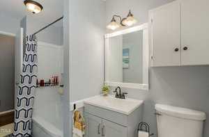 A modern bathroom with a white vanity and sink, a mirror with lights above, and a small potted plant on the counter. There's a toilet and a bathtub with a blue and white patterned shower curtain. The walls are painted light gray.