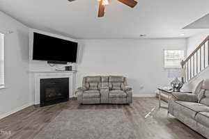 A modern living room with a ceiling fan, a wall-mounted TV above a fireplace, and two recliner sofas. There is a small side table with a lamp next to the sofas, and a window on the right. The floor is wooden with a gray rug. Stairs are visible in the back.