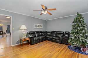 A cozy living room with a black leather sectional sofa. A decorated Christmas tree stands in the corner on a blue tree skirt. The wooden floor complements the gray walls. A ceiling fan with lights is above, and a lamp sits on a side table.