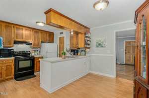 A spacious kitchen with wooden cabinets, white countertops, and light wood flooring. It features a black stove, stainless steel fridge, and a small plant on the island. Wall decor and a small chandelier add a cozy touch.