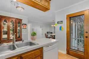 A cozy kitchen with a double sink, wooden cabinets, and a dishwasher beneath the counter. A dining area is visible, featuring a table with chairs and a china cabinet. A glass door leads to a porch with a view of outdoor greenery.