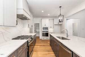 A modern kitchen with white cabinets, a marble-patterned backsplash, and a wooden floor. It features stainless steel appliances, including two ovens and a stove. A large island with a sink and pendant light completes the space.