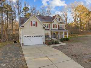 A two-story, beige house with a double garage, red shutters, and a front porch with columns. It is set in a wooded area with bare trees and a large concrete driveway, under a partly cloudy sky.