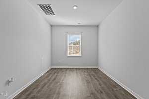 A small, empty room with light gray walls and wood flooring. There is a single window on the far wall, allowing natural light to illuminate the space. The ceiling has a recessed light fixture and a visible air vent.