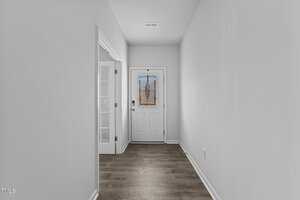 A narrow hallway with light gray walls, leading to a white front door with a decorative stained glass window. The wood floor is a medium brown, and a doorway with glass French doors is on the left side.