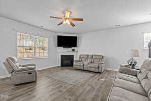 A spacious living room with a ceiling fan and hardwood floors. It features two grey sofas, a recliner, a TV mounted above a fireplace, and a lamp on a side table. Large windows with blinds allow natural light into the room.