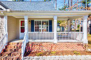 A cozy house with a front porch featuring white railings and a brick base. The porch has a red door with a window, and there's large window with blue shutters on the house. Trees and shrubs are visible in the background.