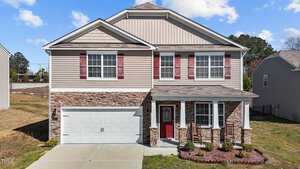 Located at 2813 E Brookwood Court, this Creedmoor two-story house boasts beige siding and stone accents. A striking red door with white-trimmed windows complements the design, and a two-car garage awaits. The landscaped front yard features lush shrubs under a clear sky dotted with a few clouds.