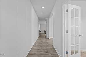A long, narrow hallway with light-colored hardwood flooring and white walls. On the right, there is a door with glass panes leading to another room. At the end of the hallway, there appears to be a sitting area with a chair.