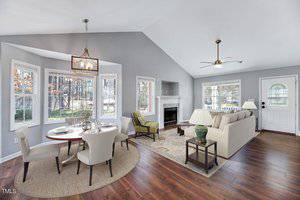 A modern living and dining area with gray walls and wood flooring. The room features a round dining table, beige chairs, a beige sofa, a fireplace, and large windows. A chandelier hangs above the dining table, and there's a ceiling fan in the living space.