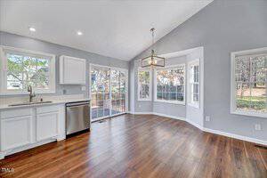 A bright, modern kitchen and dining area with wooden floors and light gray walls. It features white cabinets, a dishwasher, a window above the sink, and a large windowed door leading to a deck. A geometric pendant light hangs above.