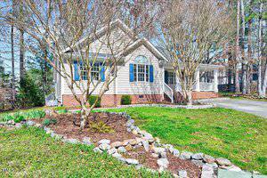 A one-story house with beige siding and blue shutters. It features a covered porch, white columns, and a brick foundation. Leafless trees are in the yard, and a stone-lined garden borders the grass. A driveway is visible to the right.