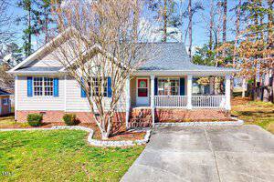 Nestled in the charming neighborhood of Creedmoor, this cozy, single-story house at 2765 Courtney Court boasts light gray siding, blue shutters, and a brick foundation. It features a welcoming covered front porch with a red door and a small tree in the yard, encircled by tall trees lining the driveway.