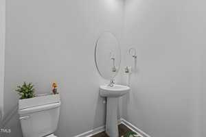 Small bathroom with a white pedestal sink and a circular mirror above it. There's a white toilet with a plant and magazines on the tank lid. The wall is painted light gray, and the floor is dark wood. A small towel ring is on the wall.