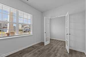 A bright, empty room with light gray walls and wood flooring. Large window with horizontal blinds on the left, allowing natural light in. Glass-paneled double doors open to another room.