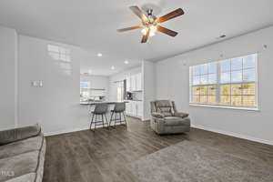 Modern living room with wood flooring, featuring a ceiling fan, recliner chair, and sofa. A small dining area with two stools is adjacent, leading to a bright kitchen with white cabinets and stainless steel appliances. Large windows allow natural light.