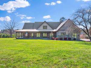 A large house with a wraparound porch and gabled roof sits on a spacious, well-manicured lawn under a blue sky. An American flag hangs by the entrance. Leafless trees are on either side of the lawn.