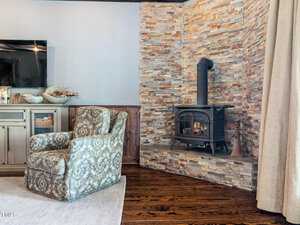 A cozy living room corner featuring a stone fireplace with a black wood-burning stove. A patterned armchair sits on a light rug, adjacent to a wooden cabinet. Neutral tones and rustic decor give the room a warm, inviting feel.