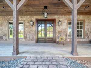 A rustic porch featuring wooden columns, a double door with glass panels, and two hanging lanterns. The brown exterior wall is textured, and a single rocking chair with a cushion sits to the side. Stone steps lead up to the porch.