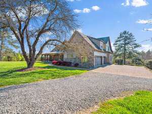 A stone house with a triangular roof sits in a sunny, grassy landscape. A gravel driveway leads to a garage attached to the side. Leafless trees and a blue sky with scattered clouds are in the background.