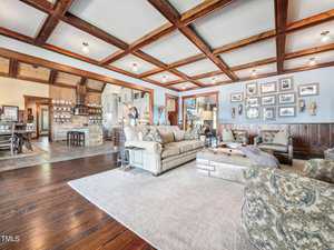 A spacious living room with wooden ceiling beams and hardwood floors. It features two sofas, a cushioned ottoman, and a light-colored rug. Various framed pictures adorn the walls, and the room leads into an adjacent dining area.