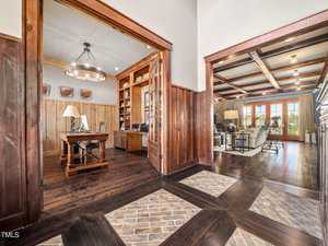 Spacious interior featuring a home office with wooden walls and desk on the left, and a cozy living room with wooden beams and large windows on the right. Warm tones and elegant decor create a welcoming atmosphere.