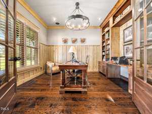 A home office featuring wooden floors and walls, a large wooden desk with a lamp, bookshelves with various decor, and a single beige armchair. A modern chandelier hangs from the ceiling, and natural light pours in through a window with blinds.
