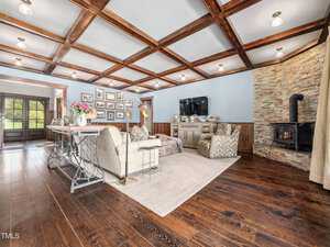 Spacious living room with wooden beam ceiling and hardwood floors. Features a stone fireplace with a black stove, a cozy seating area, a wall-mounted TV, and decorated with framed photos. A large table with flowers is near the entrance.