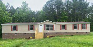 Located at 3184 Sanford Road in Oxford, this beige double-wide mobile home with a brick foundation and red shutters sits in a grassy area. Stairs lead up to the front door, and tall, lush green trees fill the background, enhancing its serene ambiance.