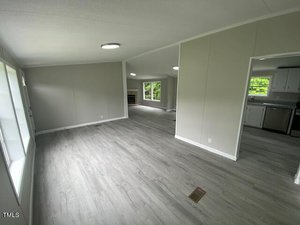 A spacious, empty room with light gray walls and wood flooring. Large windows on the left allow natural light to fill the space. In the background, there is an open doorway leading to a kitchen with visible countertops and cabinets.