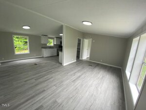 Bright, open-plan interior with light gray wood flooring. The living area flows into a modern kitchen featuring white cabinets and dark countertops. Large windows let in natural light, and the walls are painted in a soft gray tone.