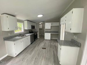 A bright, modern kitchen with white cabinets and grey granite countertops. The space includes a stove, microwave, dishwasher, and refrigerator. The flooring is light grey, complementing the neutral wall color, and a window lets in natural light.