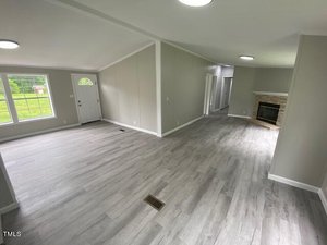 Spacious living area with light gray wood flooring, white walls, and ceiling. Includes a fireplace, multiple windows, and an angled view of the front door. Hallway leads to other rooms. Bright natural light fills the space.