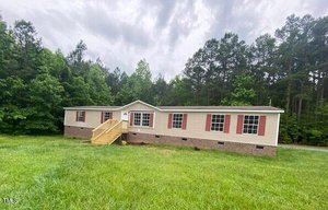 A tan manufactured home with red shutters sits on a grassy lot surrounded by trees. It features a new wooden ramp leading to the front door. The sky is overcast.