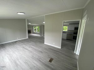 A spacious, unfurnished interior with light gray walls and flooring. The area is open-plan with a view into the kitchen, featuring white cabinetry. Large windows allow natural light to fill the space, and a doorway leads to another room.