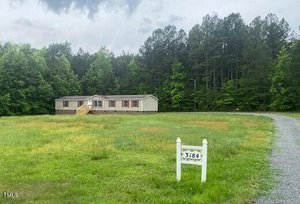 A single-story house with beige siding sits surrounded by a grassy field. A gravel driveway leads to the house. A small white sign with the number 3184 is in the foreground. Dense trees fill the background under a cloudy sky.