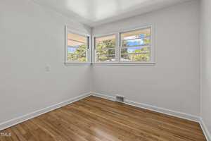 A small, empty room with light wooden flooring and white walls. Three rectangular windows on one wall let in natural light, offering a view of trees outside. The room has a clean, minimalist appearance.
