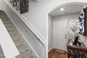 A bright entryway with a staircase on the left, leading upwards, decorated with framed photos on the wall. To the right, a wooden console table with a floral arrangement and a mirror stands against the wall by a closed white door.