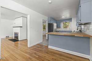 A bright kitchen with blue cabinets, wooden countertops, and white walls. It opens into a living area with a white brick fireplace. The floor is light wood, and there is a window above the kitchen sink.